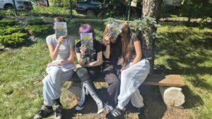 cztery osoby siedzą na ławce i zasłaniają sobie twarze książką